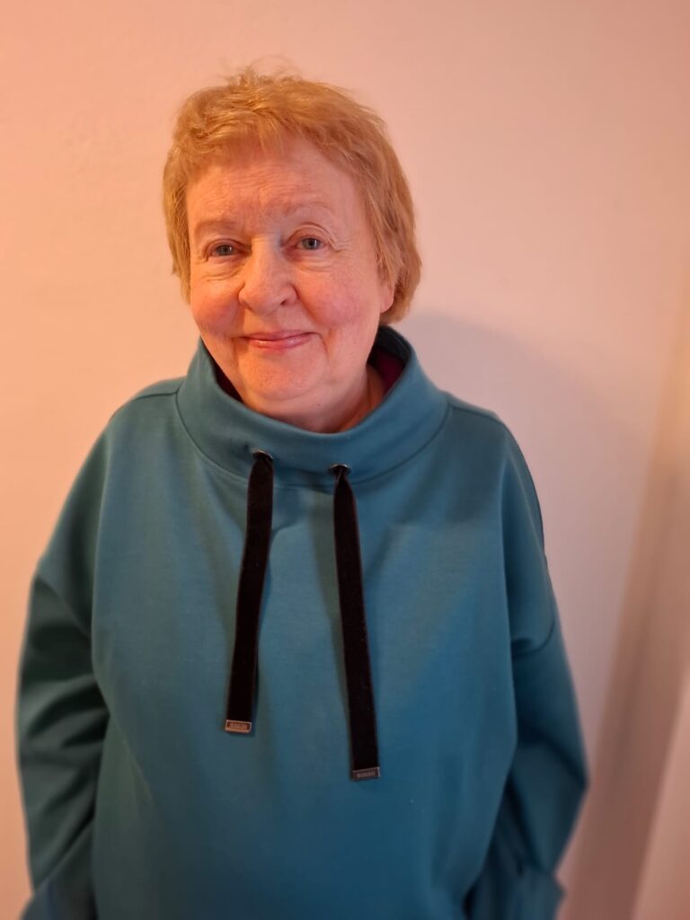 Lynne Hromek in blauem Kapuzenpulli lächelt vor heller Wand