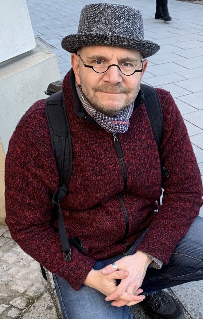 Manuel Welscher mit Hut und Brille auf Stadtpflaster sitzend
