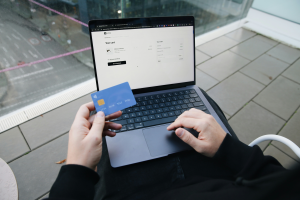 Eine Person sitzt im Freien und hält eine blaue Kreditkarte in der einen Hand, während sie mit der anderen Hand auf einem Laptop tippt, auf dem eine Online-Einkaufskassenseite angezeigt wird.